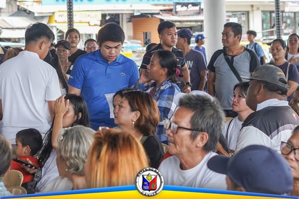 Mga tao sa Naic Town Plaza habang isinasagawa ang Medical at Burial Assistance, may lalaking nakablue na polo na may hawak…
