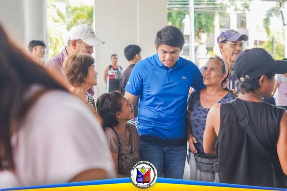Mga tao sa covered area ng Naic Town Plaza habang nakikinig sa programang Medical at Burial Assistance