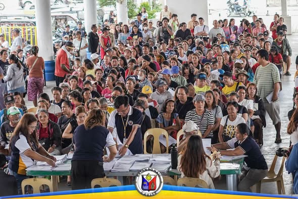 Maraming tao sa payout ng tulong sa Naic Town Plaza, habang pinoproseso ng staff ang mga dokumento sa mesa