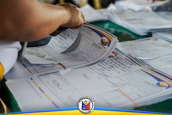 Mga nakatuping papeles at pormularyo ng ayuda na hawak ng kamay sa mesa ng payout sa Naic Town Plaza