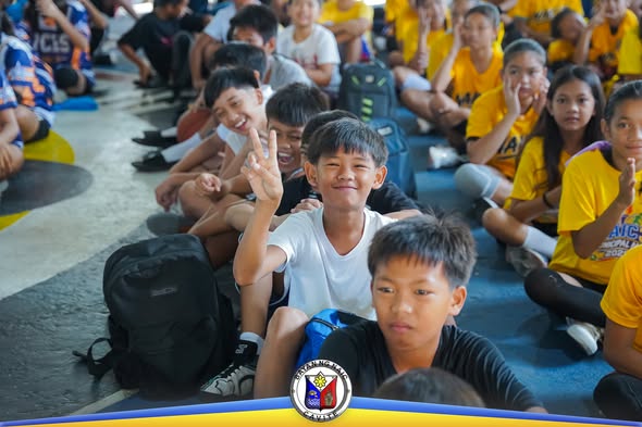 Mga batang kalahok sa Municipal Sports Meet 2026 na nakaupo sa covered court, may isang batang nagpapakita ng peace sign