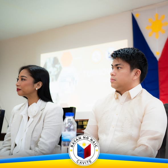Dalawang tao sa turnover ceremony ng Bayan ng Naic, nakaupo sa harap ng screen at watawat