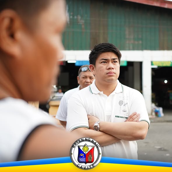 Isang opisyal na nakatayo habang naka-krus ang braso sa palengke, kausap ang mga tao sa harap
