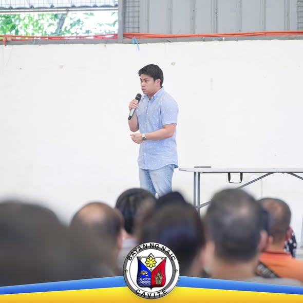 Isang lalaki na nagsasalita sa mikropono sa harap ng mga nakaupong tao sa loob ng covered court