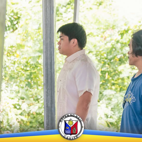 Litrato: Lalaking naka-barong sa ilalim ng open structure habang may kasamang lalaki sa gilid, may mga puno sa likod