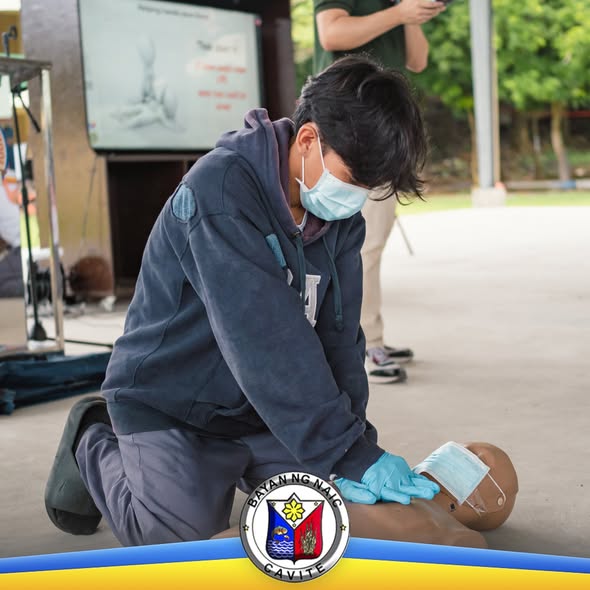 Estudyante na nagsasanay ng CPR sa manikin sa ilalim ng covered area, may suot na face mask at guwantes