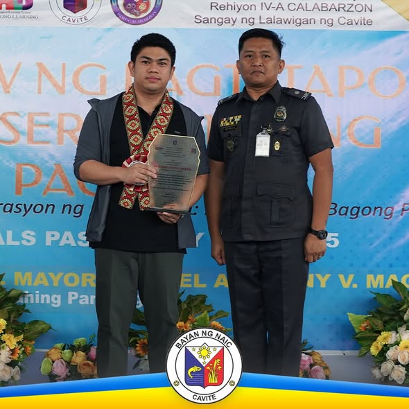 Lalaking tumatanggap ng plake habang nakatayo katabi ng opisyal na naka-uniporme sa seremonya ng pagtatapos sa Naic