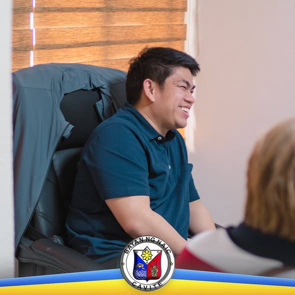 Nakangiting lalaki na nakaupo sa silya habang may kausap sa loob ng opisina