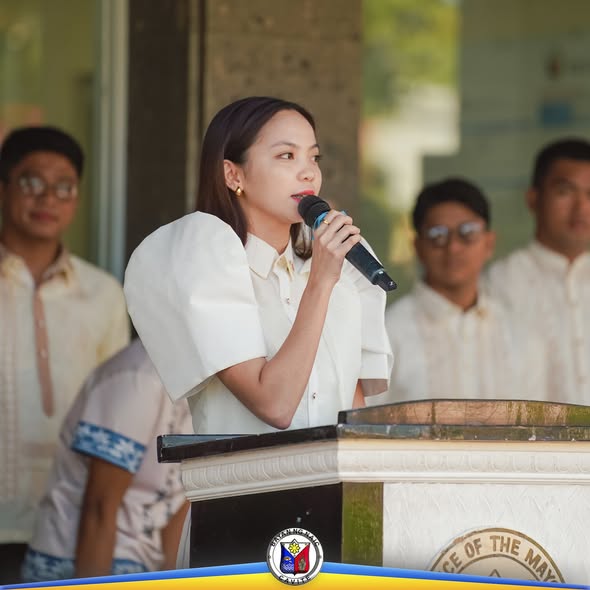Babaeng nagsasalita sa mikropono sa podium habang nakatayo sa likuran ang mga kabataan sa Flag Raising Ceremony