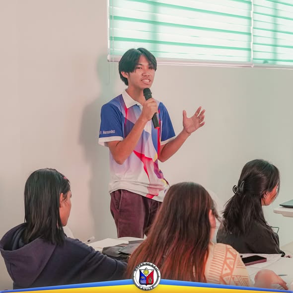 Isang kabataan na nagsasalita sa mikropono habang nakatayo sa harap ng mga kalahok sa training
