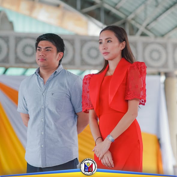 Dalawang tao na nakatayo sa event ng Buwan ng Wikang Pambansa 2025 sa Naic, may draped na dekorasyon sa likod