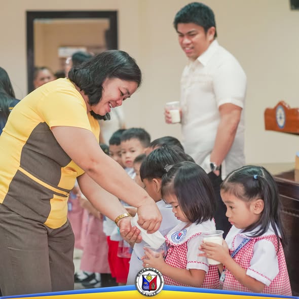Mga bata sa daycare na umiinom ng gatas habang inaalalayan ng isang kawani sa Milk Feeding Program ng Naic