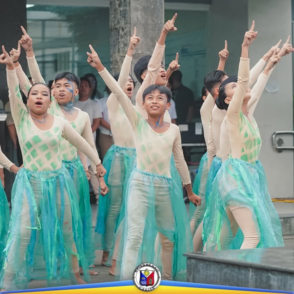 Mga estudyante na nagsasayaw-interpretasyon at nakaturo pataas sa labas ng gusali habang nagtatanghal