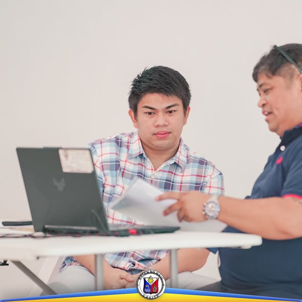 Dalawang lalaki na nag-uusap habang tinitingnan ang laptop at mga papel sa mesa sa pulong ng LDC