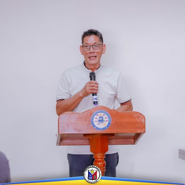 Lalaking nagsasalita sa mikropono sa podium sa harap ng puting dingding, sa pulong ng MSWM Board sa Naic