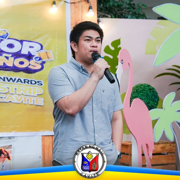 Lalaking nagsasalita sa mikropono sa harap ng banner at flamingo cutout sa job fair ng Naic