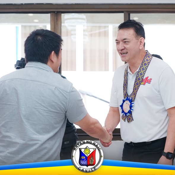 Nagkamay ang dalawang lalaki sa loob ng opisina habang nakangiti ang isa, may logo ng Bayan ng Naic Cavite