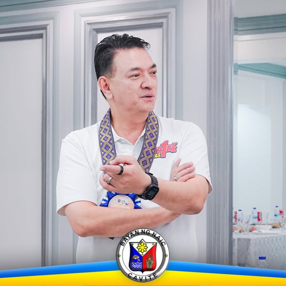 Litrato: lalaking nakahalukipkip sa loob ng silid, may lanyard at logo ng Bayan ng Naic sa ibaba
