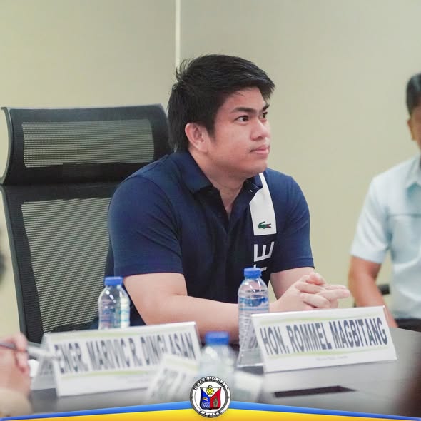 Litrato: Lalaking naka-navy polo sa pulong, may pangalan sa mesa at mga bote ng tubig