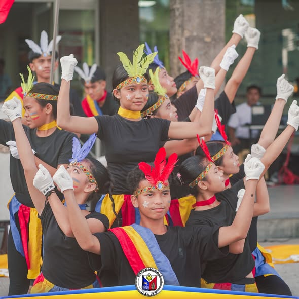 Mga mag-aaral na nagsasayaw ng interpretasyon ng Himno ng Naic sa flag raising ceremony, may balahibo at pintura sa mukha
