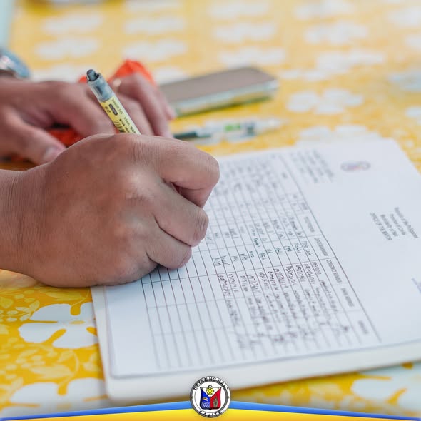 Kamay na nagsusulat sa isang talaan o form sa mesa na may dilaw na mantel sa pulong ng Local Health Board