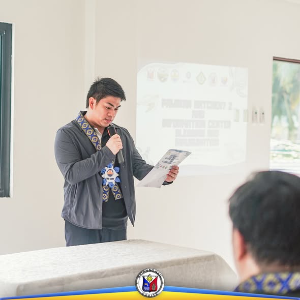 Lalaking nagsasalita gamit ang mikropono sa programa, may projected slide sa likod sa loob ng silid