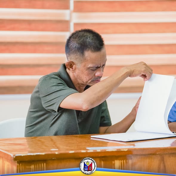 Lalaking nagbubukas ng malaking dokumento sa mesa habang nagpupulong sa Liga ng mga Barangay Office, Naic