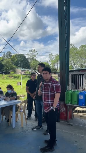 Mga kalalakihan na nakatayo at nakikinig sa harap ng mesa sa labas, sa PCSO medical mission sa Villa de Adelaida