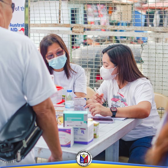 Mga kawani ng health office na nagseserbisyo sa mesa sa labas habang may mga gamot at papeles na nakalatag