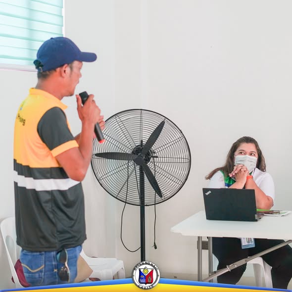 Lalaking nagsasalita gamit ang mikropono habang nakikinig ang babaeng naka-face mask sa orientation seminar
