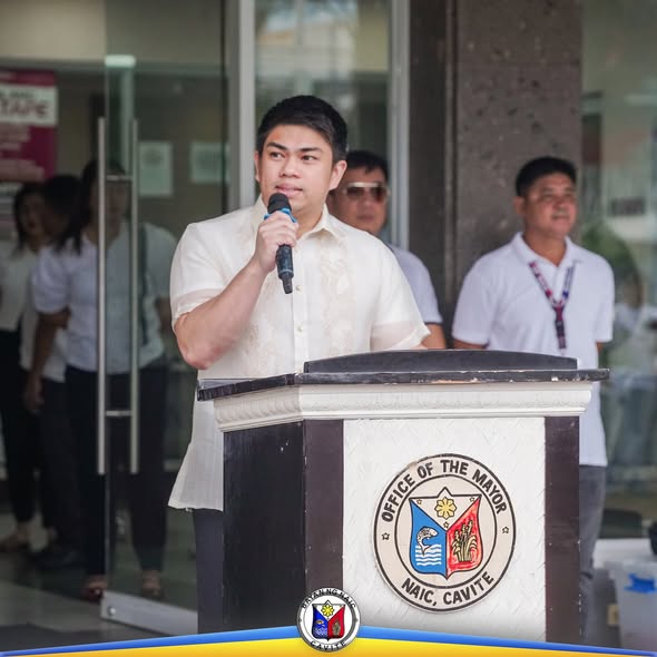 Lalaking nagsasalita sa mikropono sa podium ng Office of the Mayor, Naic, sa harap ng gusali ng munisipyo