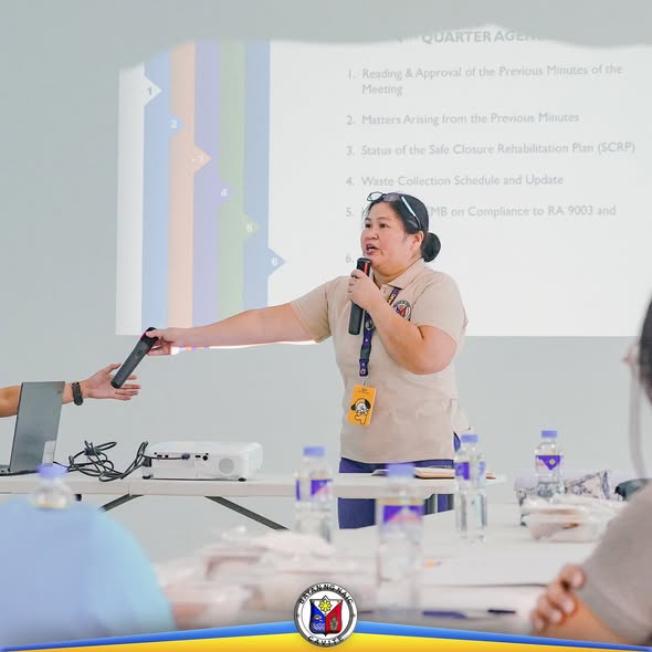 Babaeng nagsasalita sa mikropono sa pulong ng Solid Waste Management Board, may agenda sa screen sa likod