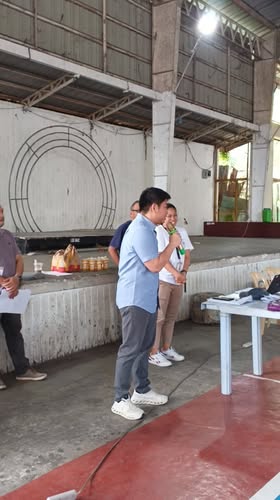 Isang lalaki na nagsasalita sa mikropono sa covered court habang may mga kasamang nakikinig sa paligid