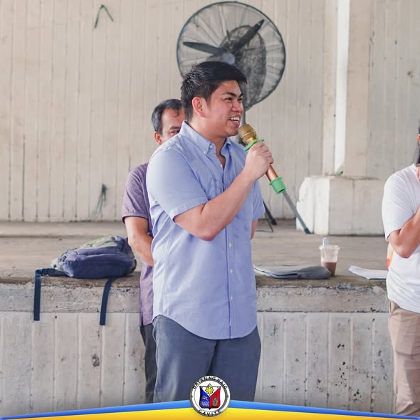 Isang lalaki na nagsasalita sa mikropono sa covered court habang may mga nakikinig sa likuran