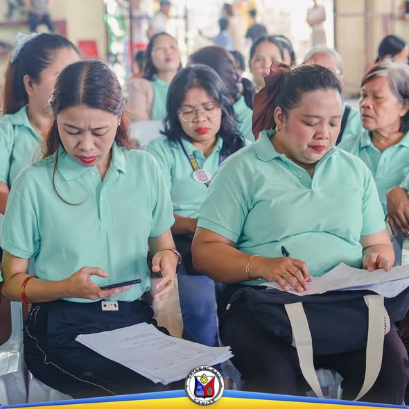 Mga benepisyaryo ng SLP na nagbabasa ng dokumento habang nakaupo sa covered court ng Naic