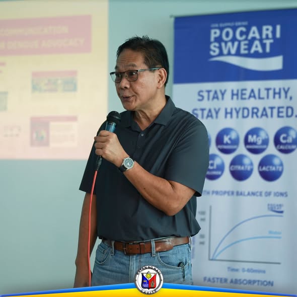 Isang opisyal na nagsasalita sa mikropono sa harap ng Pocari Sweat banner sa pagtitipon ng CAPHENA sa Naic