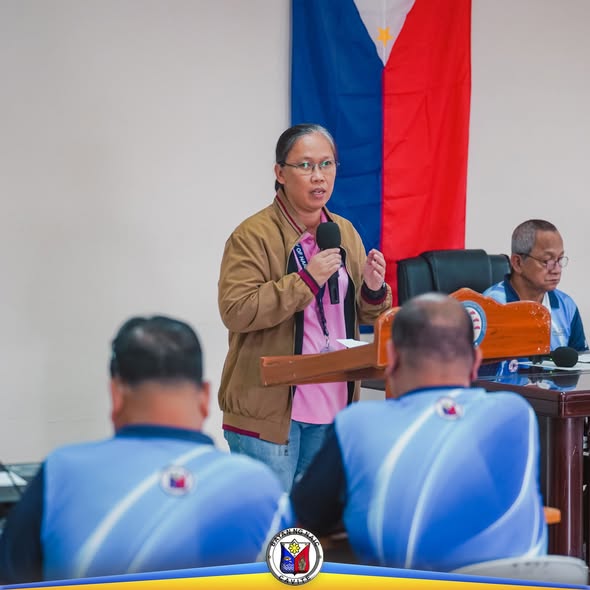 Babaeng nagsasalita sa mikropono sa pulong ng Liga ng mga Barangay sa Naic, may watawat ng Pilipinas sa likuran