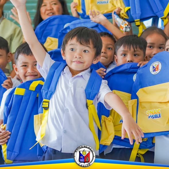 Mga batang mag-aaral na nakataas ang kamay habang hawak ang bagong school bag mula sa pamahalaang bayan ng Naic