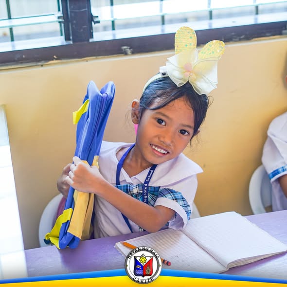 Estudyanteng nakangiti habang hawak ang asul na school bag sa loob ng silid-aralan