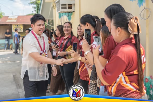 Mga kalahok na nakauniporme na kulay maroon habang nakikipagkamay sa isang lalaking naka-barong sa labas ng gusali