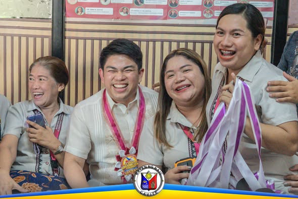 Mga masayang kalahok sa seremonya ng groundbreaking, may suot na medalya at hawak na mga ribbon sa loob ng silid