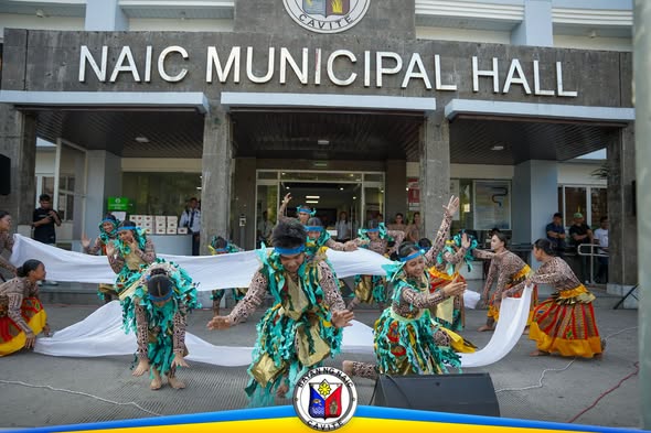 Mga mananayaw na may makukulay na kasuotan habang sumasayaw sa harap ng Naic Municipal Hall