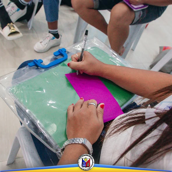 Taong nagsusulat sa papel habang nakaupo sa programang MAPANGBATA sa Naic