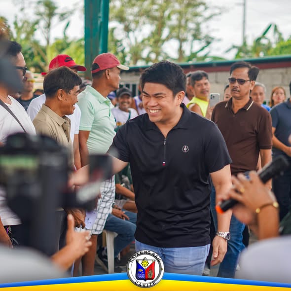 Litrato: Nakangiting opisyal na naglalakad sa gitna ng mga tao habang may mga camera at mikropono sa Brgy. Halang