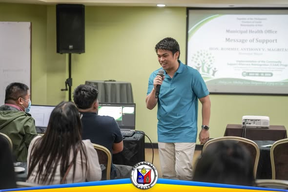 Lalaking nagsasalita gamit ang mikropono sa harap ng screen na may 'Message of Support' sa CSAR Program, Naic