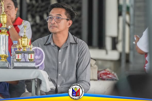 Lalaking naka-salamin na nakaupo sa programa ng Nutrition Month, katabi ng mga tropeo sa mesa
