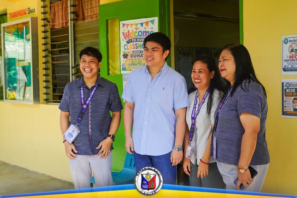 Mga opisyal at kawani ng paaralan na nakangiti sa harap ng classroom sa Halang Elementary School