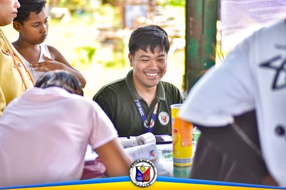 Tao na nakangiti habang nagseserbisyo sa BRAP sa Brgy. Halang, may mesa at mga papeles