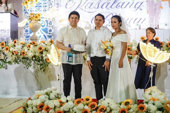 Mga kasal na mag-asawa at mga bisita sa entablado ng Kasalan ng Bayan 2026 na may puting at orange na bulaklak