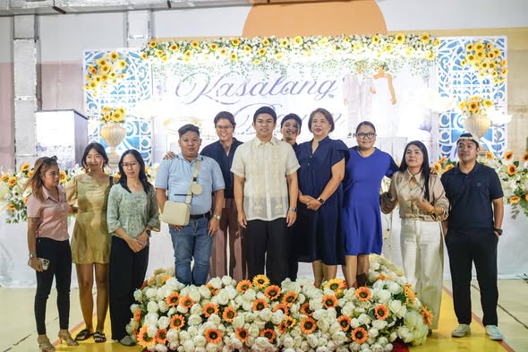 Grupo ng mga tao na nakapose sa harap ng entablado ng Kasalan ng Bayan 2026 na may bulaklak na dekorasyon